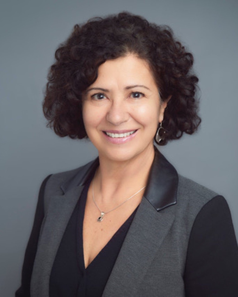 Portrait of Gloria, smiling, in a grey jacket against a grey backdrop